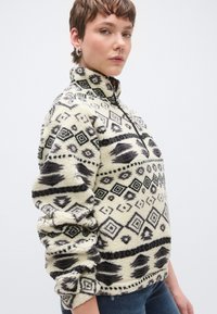 Pulover od flisa u krem boji s crnim geometrijskim uzorcima. Ima visok ovratnik i patentni zatvarač, teksturiran, lagano prevelik kroj s nabranim rukavima.