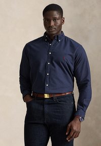 Mann iført en marineblå knappeskjorte, mørke jeans, brunt belte og klokke, stående med en hånd i lommen mot en ensfarget bakgrunn.