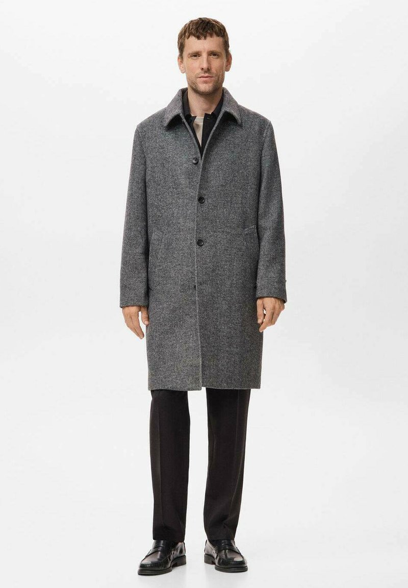 Grå ullkappa med slagkrage, knäppning, sidofickor och medellång design, kombinerad med svarta byxor och skor.