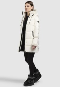Vit vadderad parka med huva fodrad med fuskpäls, dragkedja och tryckknappar, två sidofickor och svarta detaljer. Bärs över svarta leggings och stövlar.