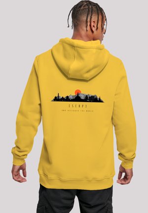 Mann mit geflochtenem Haar, der einen gelben Hoodie mit schwarzer und grauer Stadtsilhouette und Berggrafik sowie einer orangefarbenen Sonne auf dem Rücken trägt, steht vor einem grauen Hintergrund.