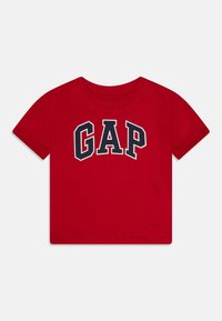Sarkans kokvilnas t-krekls ar īsām piedurknēm, uz kura krūtīm ir boldā, arku formā izdrukāts tumši zils "GAP" logo.