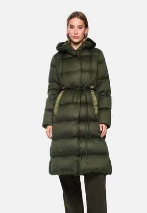 Woman wearing a long, quilted, dark green puffer coat with a hood and waist tie, standing against a plain light background.