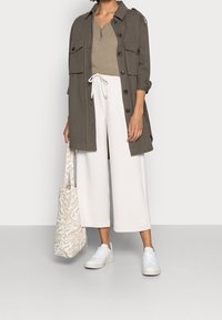 Giacca verde oliva con grandi tasche sopra una maglia beige, abbinata a pantaloni bianchi a gamba larga e sneakers bianche. Borsa a spalla beige con motivo.