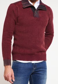 Burgunderfarbener Strickpullover aus strukturiertem Gewebe, mit einem halben Reißverschluss-Kragen, der einen kontrastierenden dunkelbraunen Akzent aufweist, sowie gerippten Bündchen und Saum.