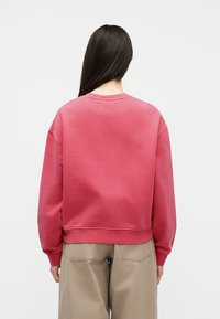Femme aux cheveux longs et foncés portant un sweat-shirt rouge ample et un pantalon beige, debout de dos sur un fond clair uni.