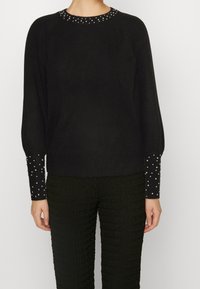 Suéter negro con un acabado texturizado, con un cuello redondo adornado con tachuelas plateadas y mangas largas con detalles de lunares.