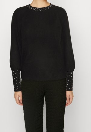 Un pull noir avec une finition texturée, doté d'un col rond orné de clous argentés et de manches longues avec des détails à pois.