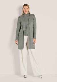 Vrouw gekleed in een grijze gebreide cardigan en sweater met een witte wijde broek en metallic puntige schoenen, staande tegen een eenvoudige achtergrond.