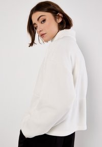 Hvid fleece-pullover med høj krave, afslappet pasform og sidelommer. Blød tekstur og minimalt søm fremhæver designet.