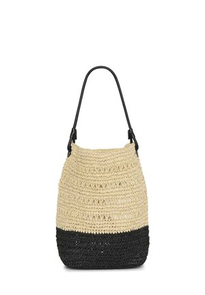 Femme portant une combinaison beige sans manches avec une taille nouée, tenant un grand sac en tissu tissé bicolore et portant des sandales noires, debout devant un fond blanc.