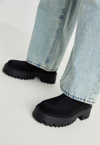 Černé platformové boty s robustní, texturovanou podrážkou, potažené hladkým materiálem. Nosené s světlými modrými volnými džíny.