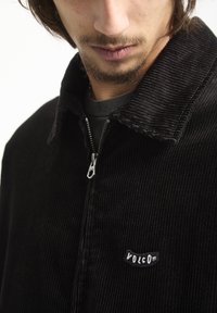 Zwart corduroy jack met een rits aan de voorkant, kraag en een Volcom-logo patch op de borst. Getextureerde stof en relaxte pasvorm.