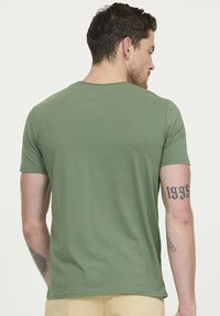 Homme aux cheveux courts et noirs portant un t-shirt vert et un pantalon beige, vu de dos, montrant des tatouages sur les deux bras.