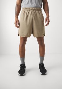 Pantalones cortos deportivos beige con cinturilla elástica y aberturas laterales, combinados con zapatillas negras y calcetines grises. Presenta el logo de Nike.
