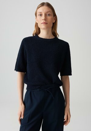 Frau mit hellbraunen Haaren, die einen strukturierten schwarzen Kurzarmpullover und schwarze Hosen trägt, steht vor einem einfachen hellen Hintergrund.