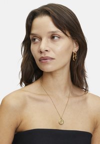 Collana d'oro con pendente a goccia, abbinata a orecchini a cerchio d'oro. Il modello indossa un top senza spalline, mettendo in risalto il design della gioielleria.