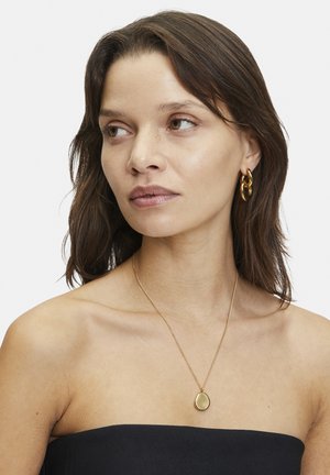 Gouden ketting met een traanvormige hanger, gecombineerd met gouden oorbellen met een ring. Model draagt een strapless top, waarmee het ontwerp van de sieraden wordt getoond.