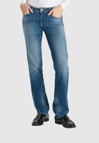 Jeans en denim bleu clair avec une coupe droite, des surpiqûres contrastantes et une fermeture à bouton métallique, associés à des chaussures noires.