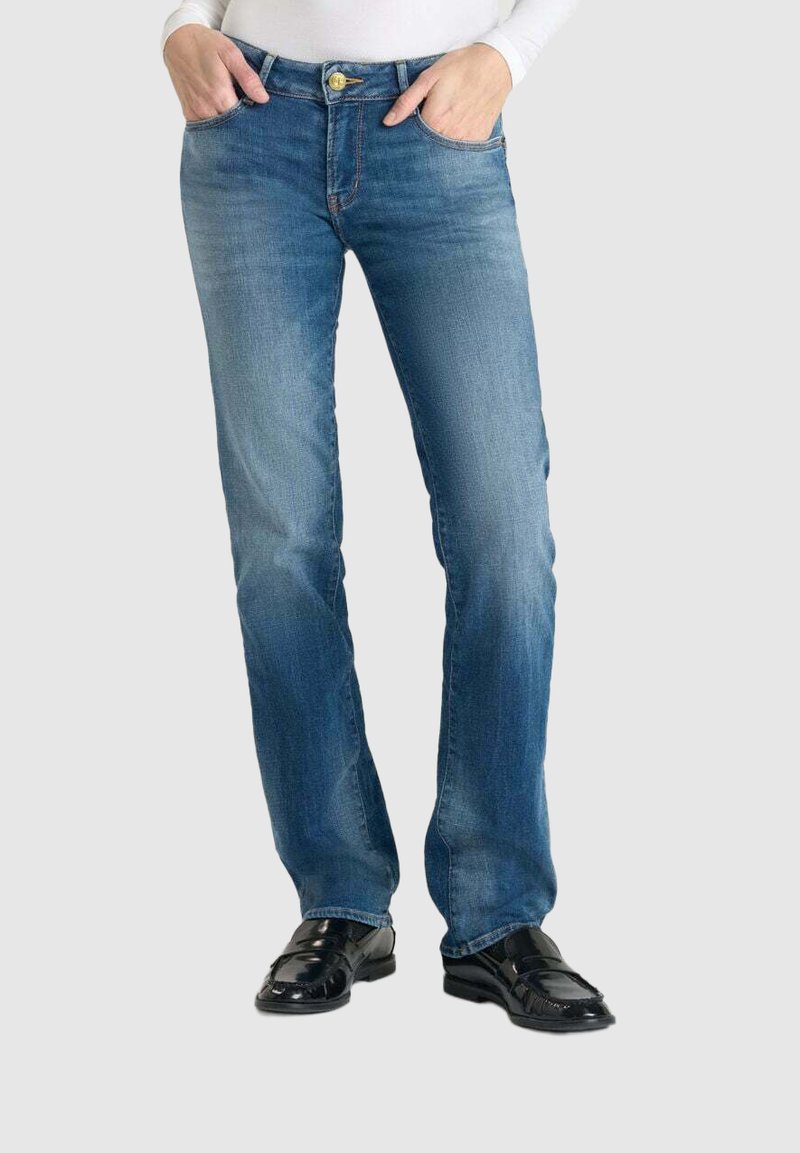 Jeans en denim bleu clair avec une coupe droite, des surpiqûres contrastantes et une fermeture à bouton métallique, associés à des chaussures noires.