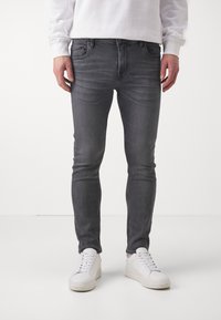 Jeans de mezclilla gris de corte ajustado, con un ligero desgastado y costuras en contraste, combinados con zapatillas blancas sobre un fondo neutro.