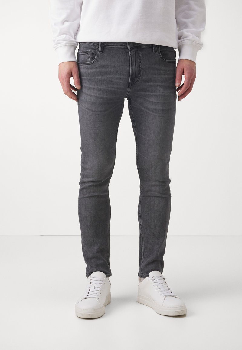 Jeans de mezclilla gris de corte ajustado, con un ligero desgastado y costuras en contraste, combinados con zapatillas blancas sobre un fondo neutro.