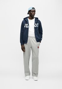 Ναυτικό μπλε φούτερ με φερμουάρ και λευκό λογότυπο, συνδυασμένο με ανοιχτό γκρι φόρμες με μικρό ετικετακι λογοτύπου; τοποθετημένο σε απλό φόντο.