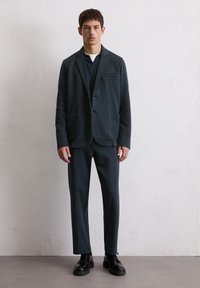 Donkergroene blazer met twee voorzakken, gecombineerd met bijpassende broek en zwarte veterschoenen; gladde stof, moderne snit en op maat gemaakt ontwerp.