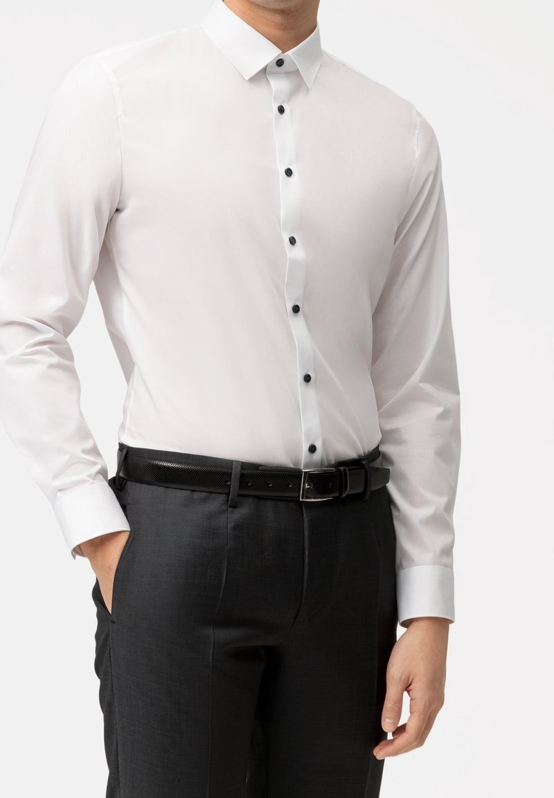 Homme portant une chemise blanche ajustée avec des boutons noirs, rentrée dans un pantalon gris foncé avec une ceinture noire, debout avec une main dans la poche.