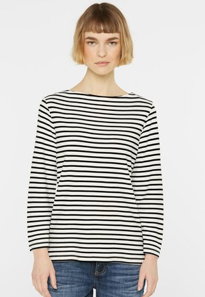 Junge Frau mit kurzen blonden Haaren, die ein langärmliges schwarz-weiß gestreiftes Shirt und blaue Jeans trägt, schaut nach vorne.