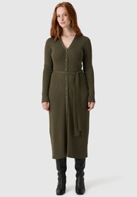 Olivegrünes, geripptes Langarmkleid mit V-Ausschnitt, Knöpfen vorne und gebundenem Taillenband. Getragen mit schwarzen kniehohen Stiefeln. Glatte Textur.