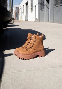 Paire de bottes de randonnée à plateforme en daim marron placée sur un trottoir urbain ensoleillé près d'une voiture garée et d'immeubles industriels.