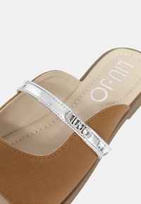 Infradito color tan con una parte superiore testurizzata, una fascia argentata lucida con branding e una soletta beige liscia.