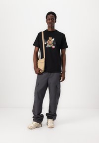 Zwart grafisch T-shirt met een beerontwerp, gecombineerd met grijze cargobroeken en beige schoenen, met een kleine beige schoudertas.