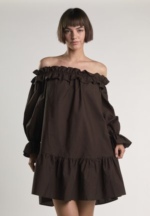Frau mit kurzen braunen Haaren, die ein schulterfreies dunkelbraunes Kleid mit Rüschen an Ärmeln und Saum trägt und vor einem schlichten Hintergrund steht.