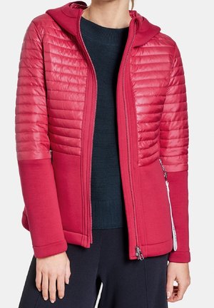 Kvinde iført en rød quiltet jakke med lynlåsen delvist åben, som viser en mørkeblå sweater under, stående mod en hvid baggrund.