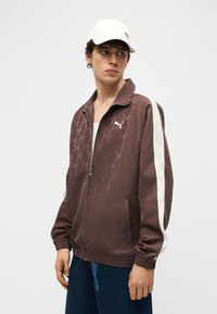 Νέος άνδρας με καφέ μπουφάν Puma με φερμουάρ και λευκές ρίγες στα μανίκια και λευκό καπέλο Puma, κοιτάζει προς τα πλάγια.