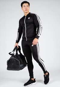 Veste et pantalon de sport noirs avec des rayures blanches sur les côtés, dotés d'une fermeture éclair à l'avant, d'une coupe ajustée et d'un logo. Tenant un sac de sport noir.