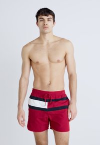 Rote Badeshorts mit schwarz-weißem horizontalem Streifenmuster, ausgestattet mit einem elastischen Bund und Kordelzug. Hergestellt aus leichtem Material.
