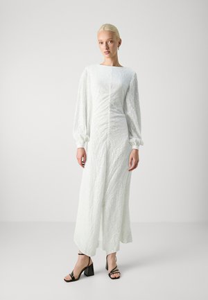 Robe blanche à manches longues avec une surface texturée et des manches bouffantes. Corsage ajusté s'évasant en une jupe fluide. Sandales à talons noires avec lanières.