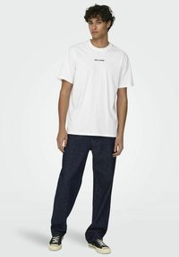 T-shirt bianco in cotone con maniche corte, caratterizzata da un logo stampato nero "ONLY & SONS" sul petto. Abbinata a jeans di denim blu scuro.