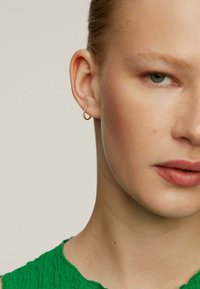 Pendiente de aro dorado con un acabado texturizado, colocado en la oreja izquierda, acentuado por pequeñas piedras transparentes. Prenda verde texturizada visible.