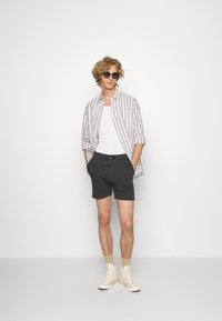 Lichte gestreepte button-up blouse in beige en wit, gedragen over een witte tanktop, zwarte shorts en crèmewitte sneakers. Eenvoudige accessoires zoals een zonnebril.