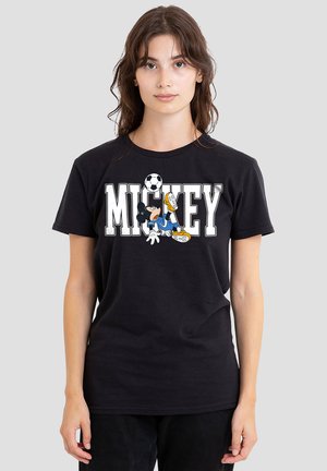Young woman wearing black T-shirt with "MICKEY" text and Mickey Mouse playing soccer graphic, standing against a plain light background.