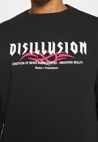 El sudadera negra presenta un audaz texto gráfico en blanco y rosa. El diseño incluye las frases "DESILUSIÓN" y "CONDICIÓN DE ESTAR DESENCANTADO".