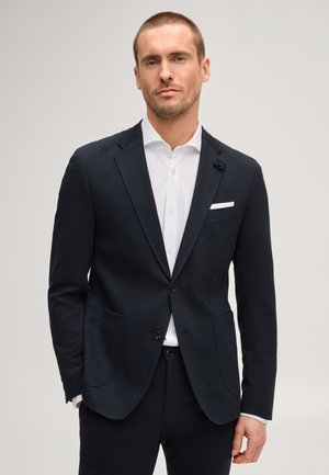 Man draagt een zwart colbert en wit overhemd, staat voor een effen lichte achtergrond, handen ontspannen langs zijn zij.