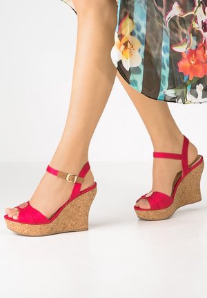 Sandales compensées avec talons en liège, brides en tissu rouge, fermeture à boucle à la cheville, portées avec une jupe à motif floral pieds nus.