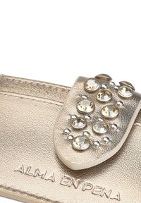 Cartera texturizada dorada con una solapa que presenta piedras decorativas transparentes y doradas claras, además de acentos metálicos. Logo grabado en la parte inferior.