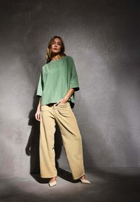 Haut oversized vert à manches courtes associé à un pantalon large en velours côtelé beige et des chaussures pointues de couleur claire, sur un fond gris texturé.