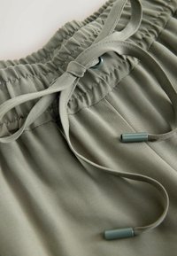 Shorts en tissu vert clair avec taille élastiquée, dotés d'un cordon de serrage avec embouts verts foncés. Texture lisse avec des rayures subtiles visibles.